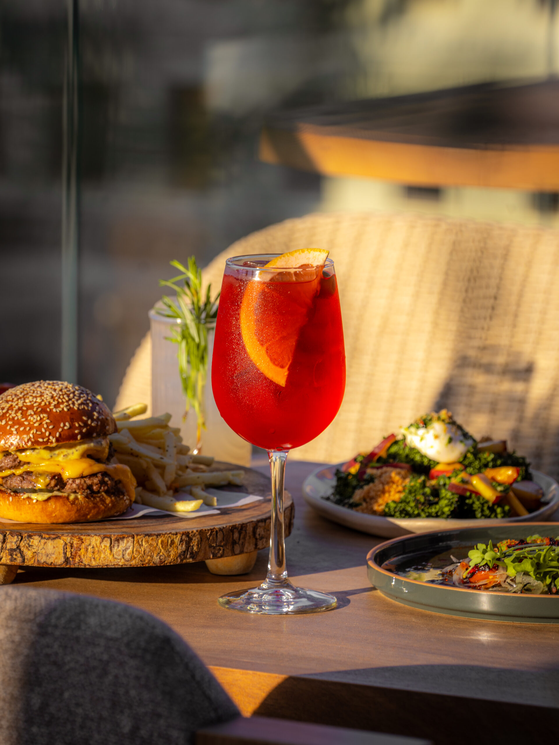 Red cocktail garnished with an orange slice served alongside a cheeseburger with fries and plated dishes on a restaurant table