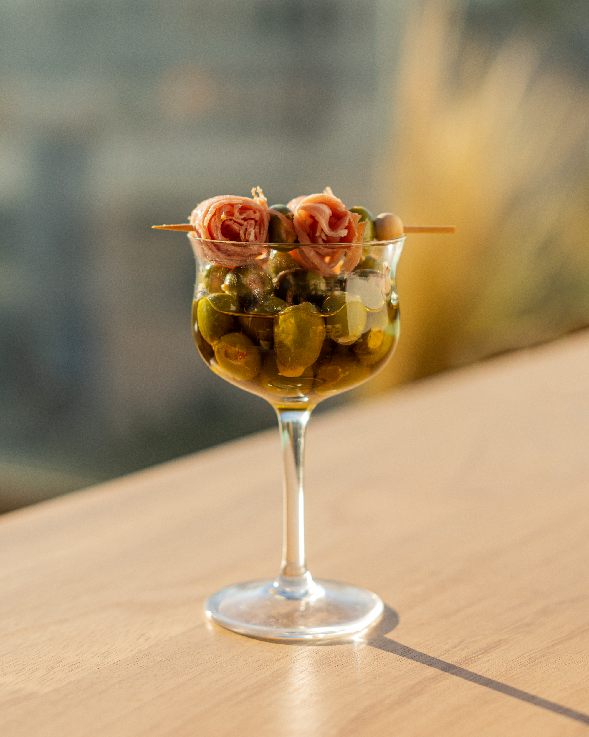 Marinated olives served in a stemmed glass topped with prosciutto and cocktail skewers