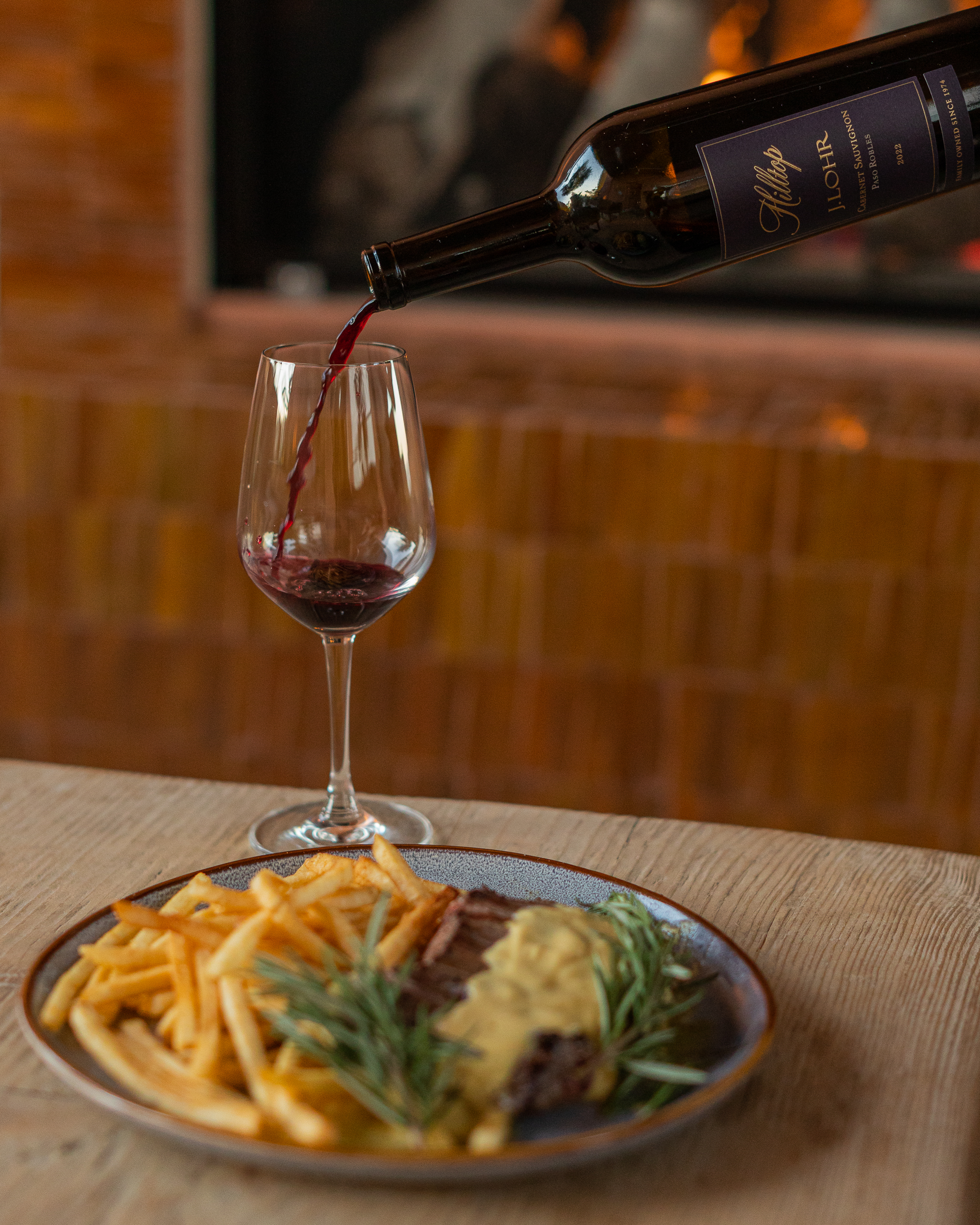 Red wine being poured into a glass beside a plate of steak with fries and sauce on a wooden table