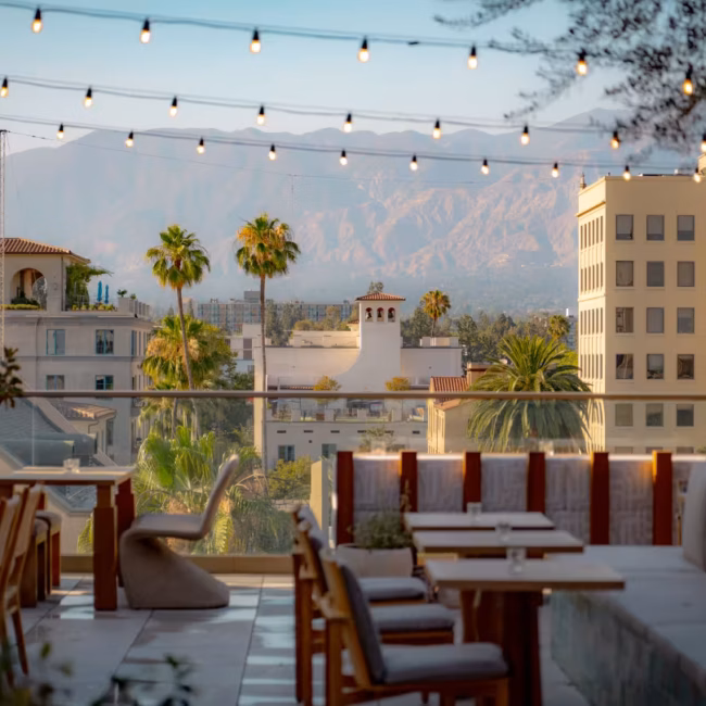 Rooftop bar seating with wooden stools and a counter overlooking city buildings and mountains at sunset