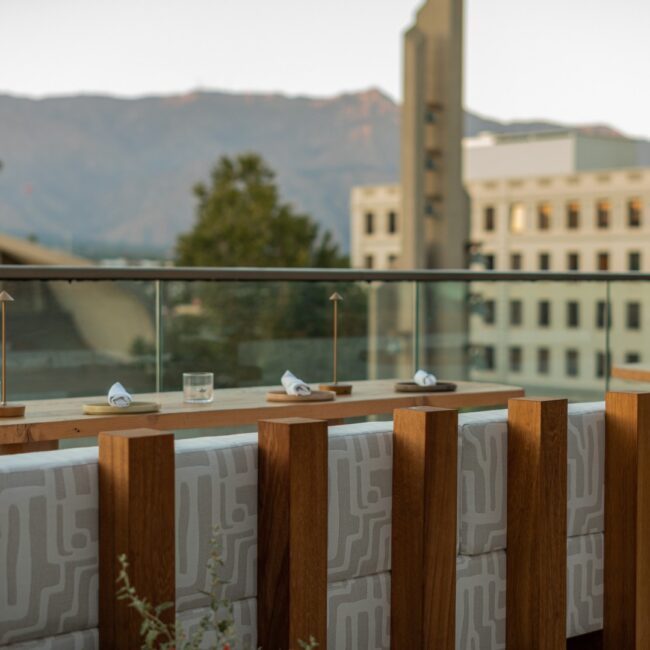Rooftop bar seating with wooden stools and a counter overlooking city buildings and mountains at sunset